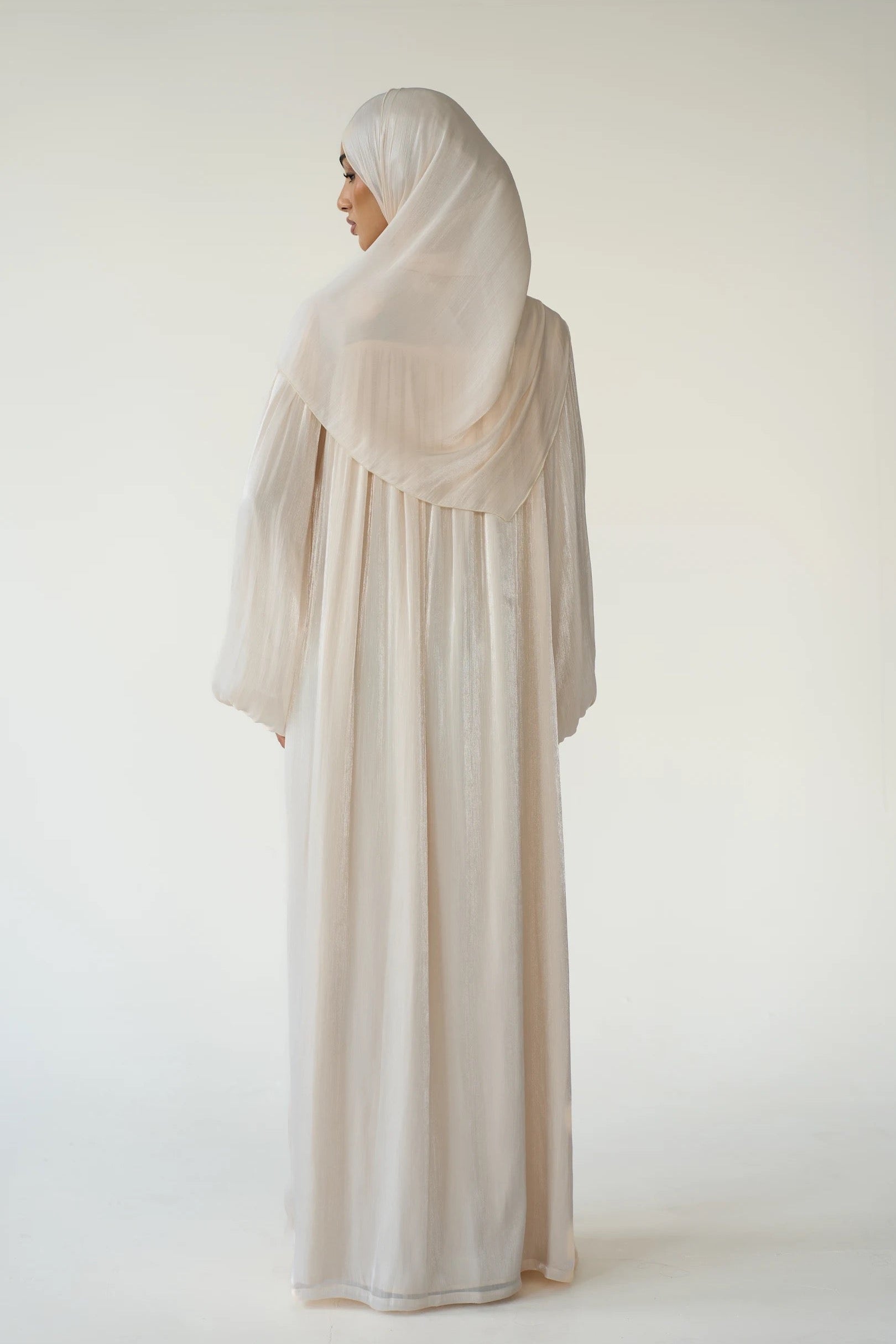 A woman wearing a flowing, lightweight abaya dress and matching hijab in a soft off-white or cream color, shown from the back against a plain, light studio background.