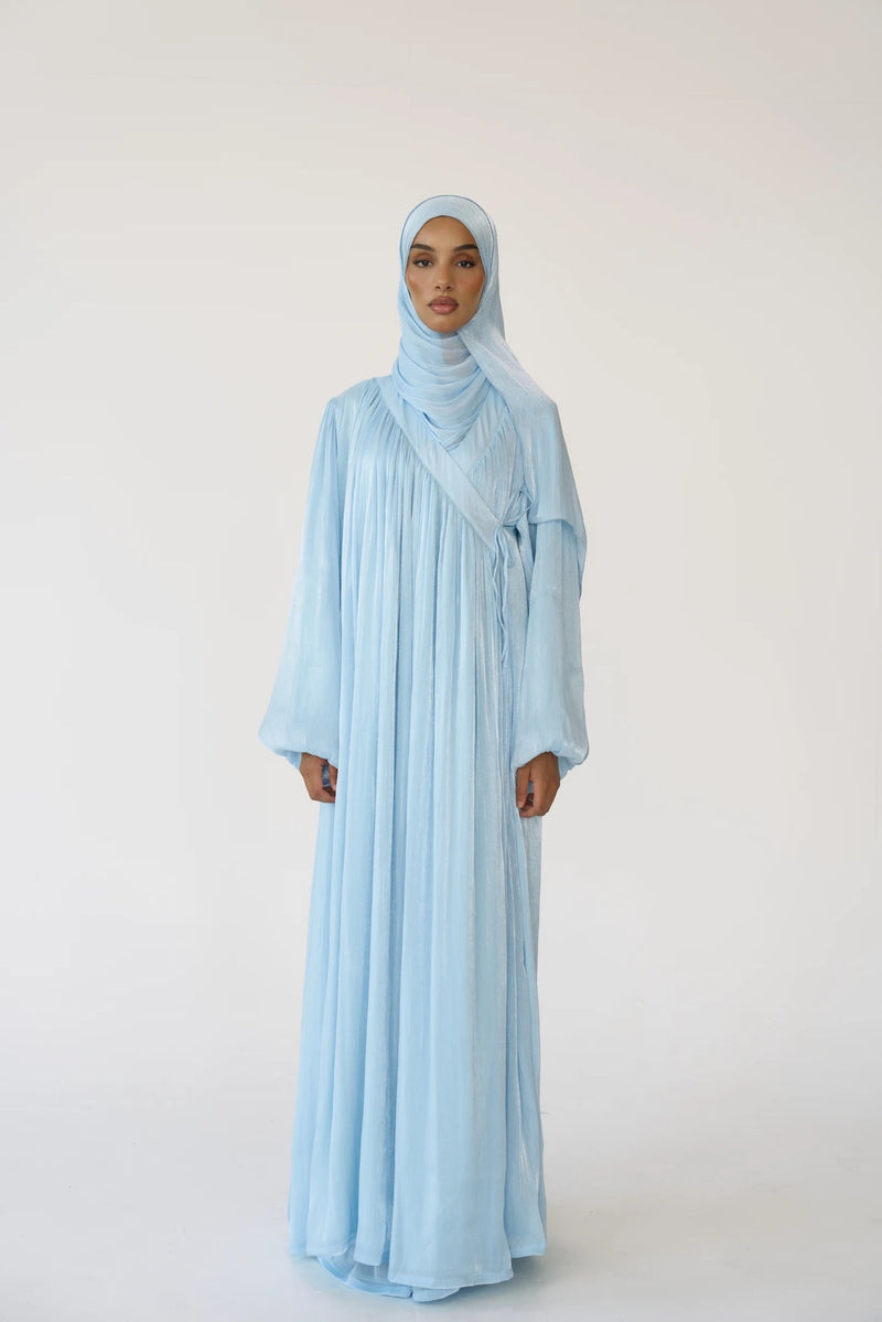 A woman modeling a light blue modest outfit consisting of a long flowing abaya with wide sleeves and a matching hijab, standing against a plain light background.